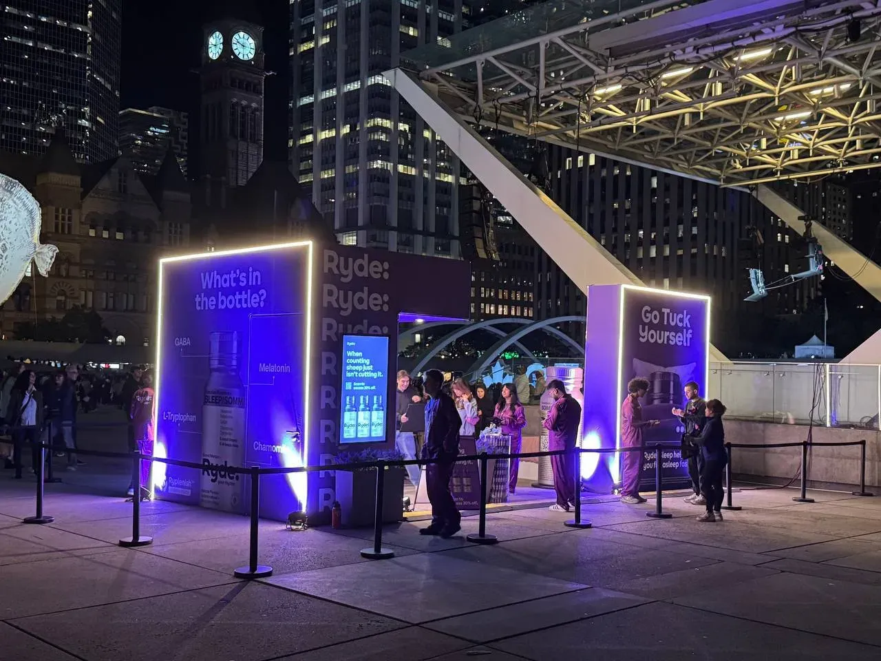 Black belt stanchions with black belts roping off the queue at a lit outdoor brand booth at night